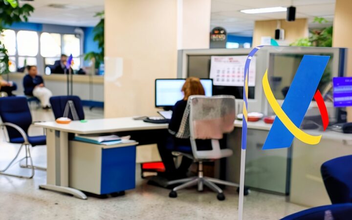 Oficina de la Agencia Tributaria con un logotipo azul y amarillo en primer plano sobre un separador de cristal transparente y empleados trabajando al fondo.