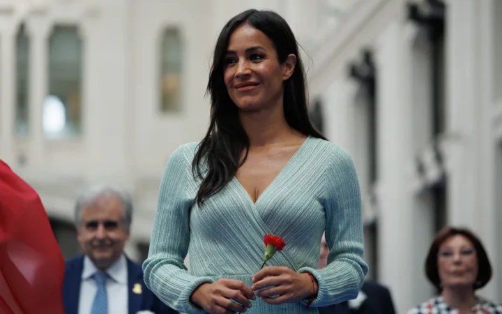 Fotografía de Begoña Villacís sonriendo sosteniendo un clavel rojo con personas al fondo.