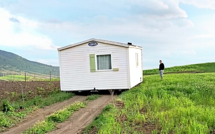 Fotografía editorial de una casa móvil blanca instalada en una parcela rústica con un camino de tierra y una persona de pie en el campo verde.