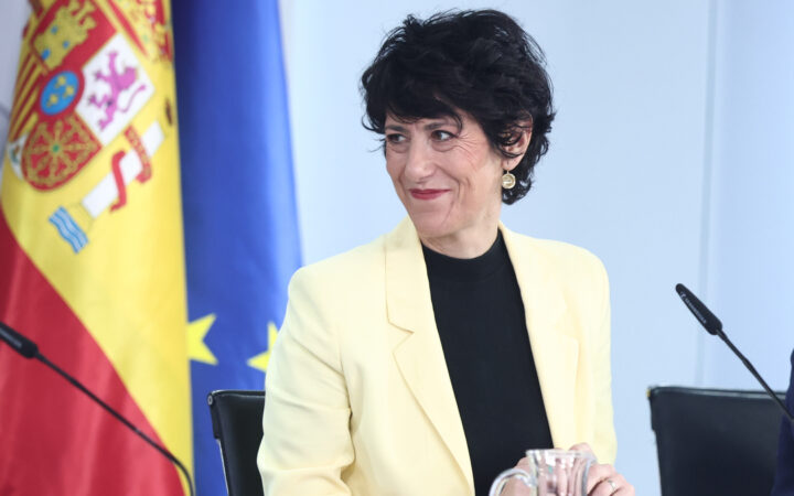 Fotografía editorial de la ministra Elma Saiz sonriendo ante un atril con la bandera de España y la Unión Europea de fondo.