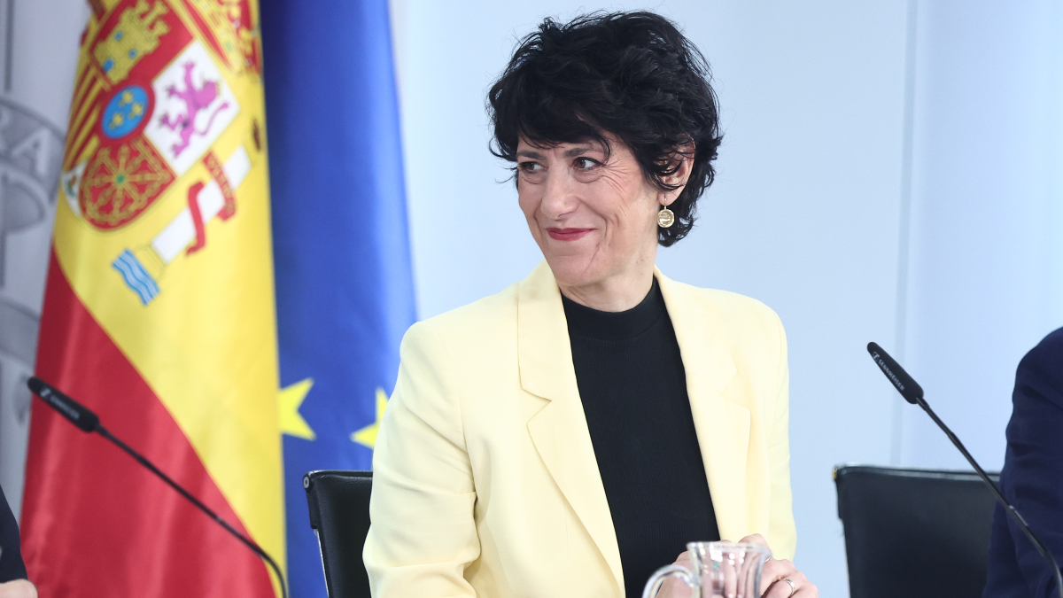 Fotografía editorial de la ministra Elma Saiz sonriendo ante un atril con la bandera de España y la Unión Europea de fondo.