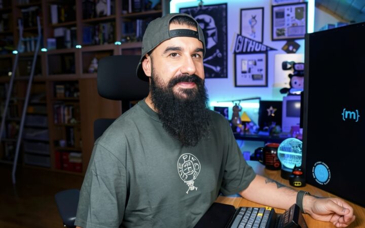 Fotografía editorial de Brais Moure sonriendo con barba poblada y gorra verde en su espacio de trabajo tecnológico.